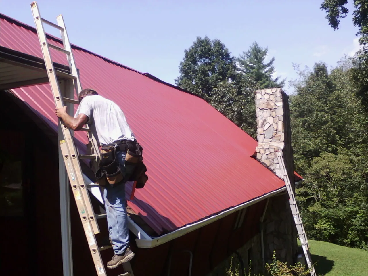 Roofer on a ladder beside a red metal roof and stone chimney — Soffit & Fascia Repair by Roofing Brothers in East Hartford